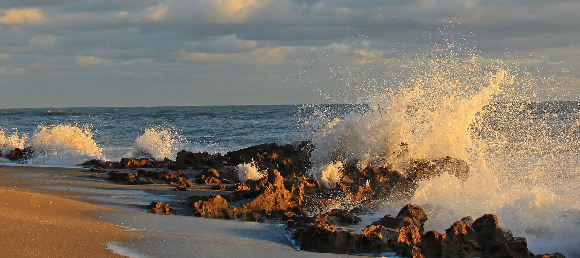 Waves crashing against rocky beach at sunset with cloudy sky.
